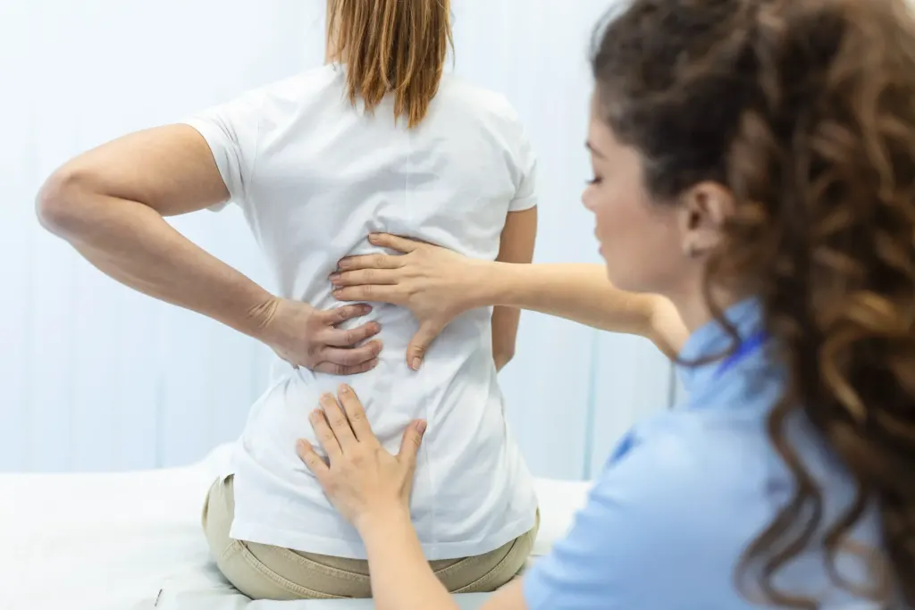 Mulher está em uma consulta com ortopedista e coloca as mãos nas costas com dores de escoliose.